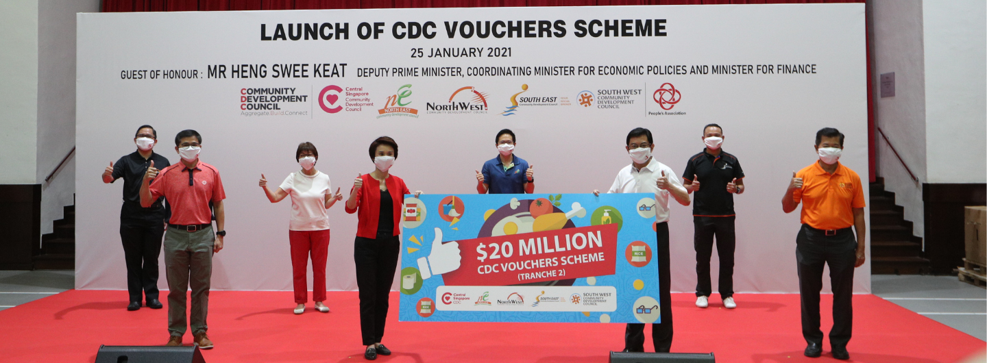 Group poses with "$20 Million CDC Vouchers Scheme" banner, giving thumbs up, wearing masks.